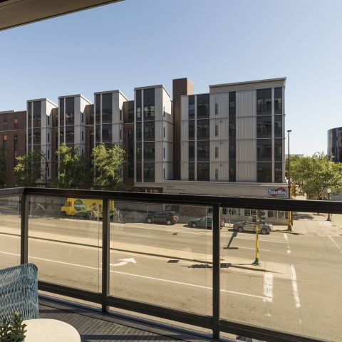 View from a balcony showcasing modern apartment buildings, a street below, and clear blue skies. A cozy chair with a decorative cushion is visible.
