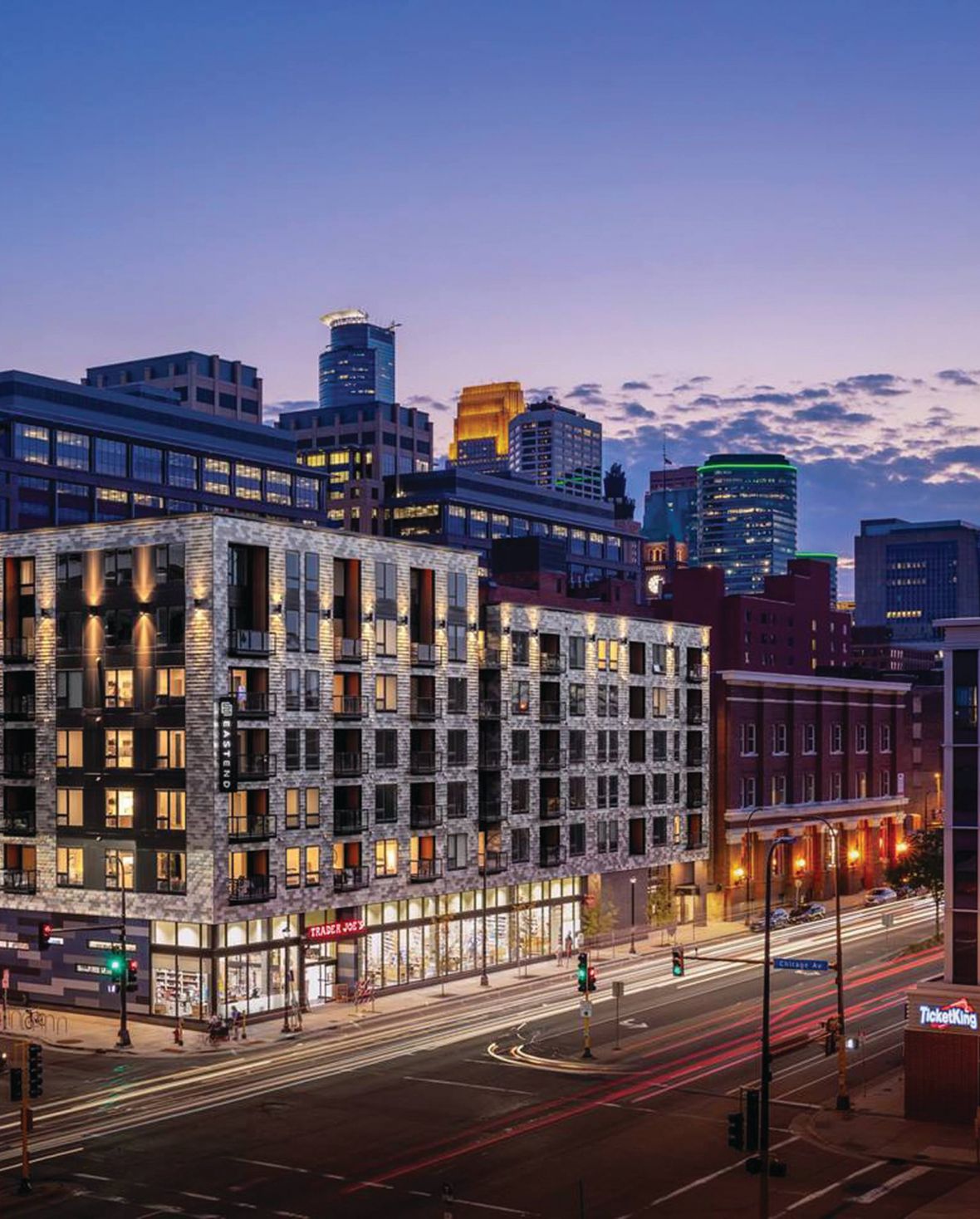 Modern urban building with illuminated apartments, bustling street below, and a vibrant city skyline at dusk.