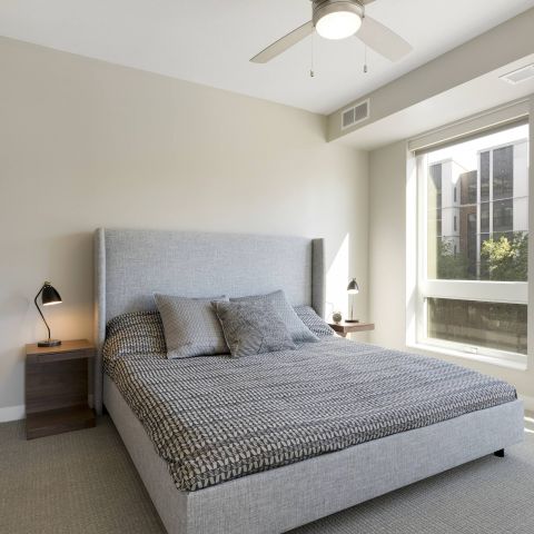Modern bedroom featuring a gray bed with patterned bedding, nightstand, lamp, and large windows allowing natural light in.