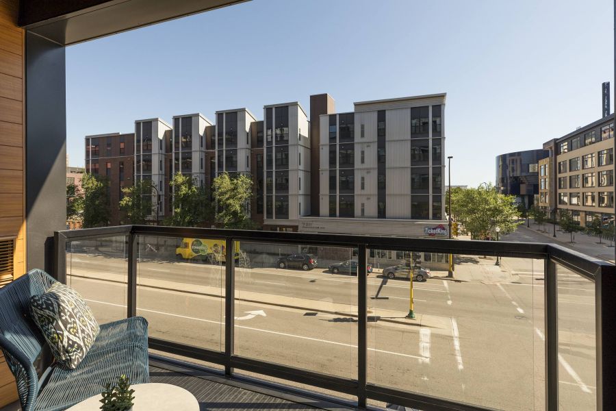 View from a balcony showcasing modern apartment buildings, a street below, and clear blue skies. A cozy chair with a decorative cushion is visible.